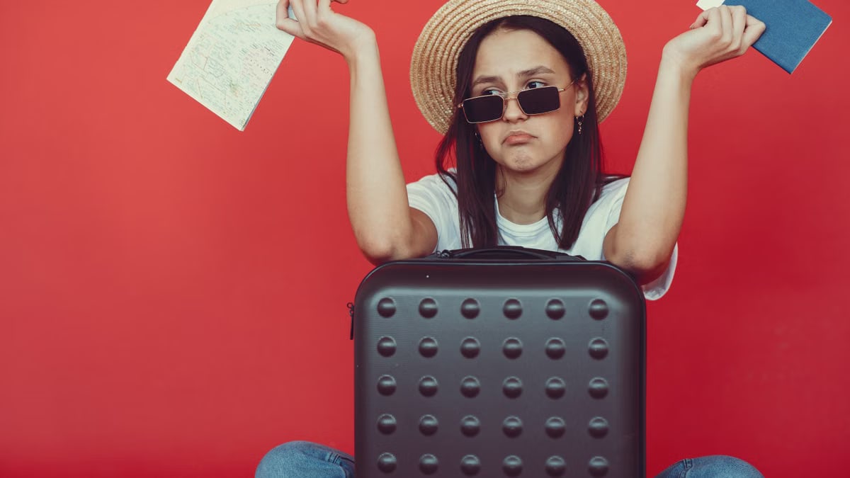 Doubters young stylish woman in wicker hat and trendy sunglasses sitting behind black suitcase and shrugging with passport and tickets on red background